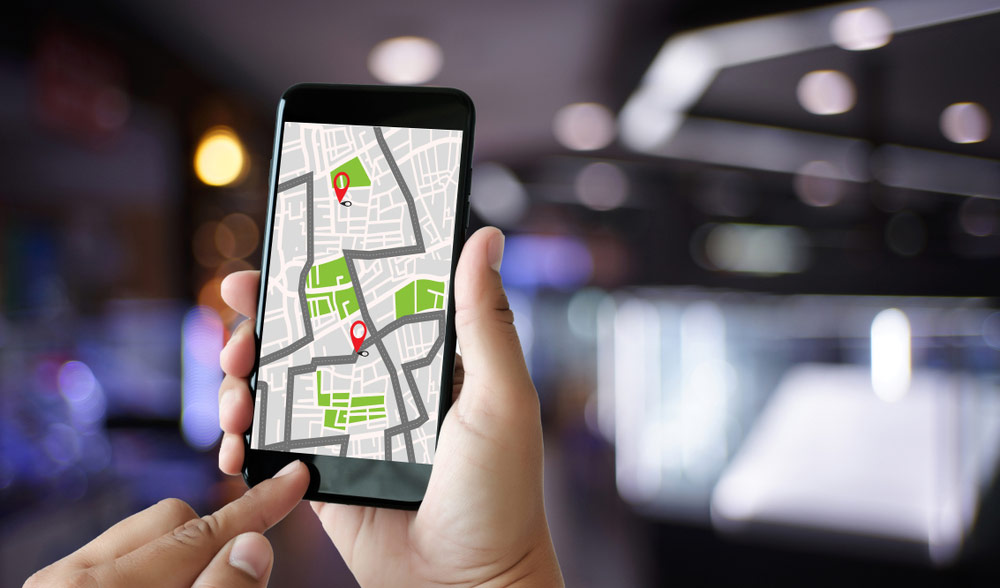 Widok ręki trzymającej smartfona z otwartą aplikacją GPS, na której widoczny jest punkt docelowy oznaczony pinezką GeoNAVI